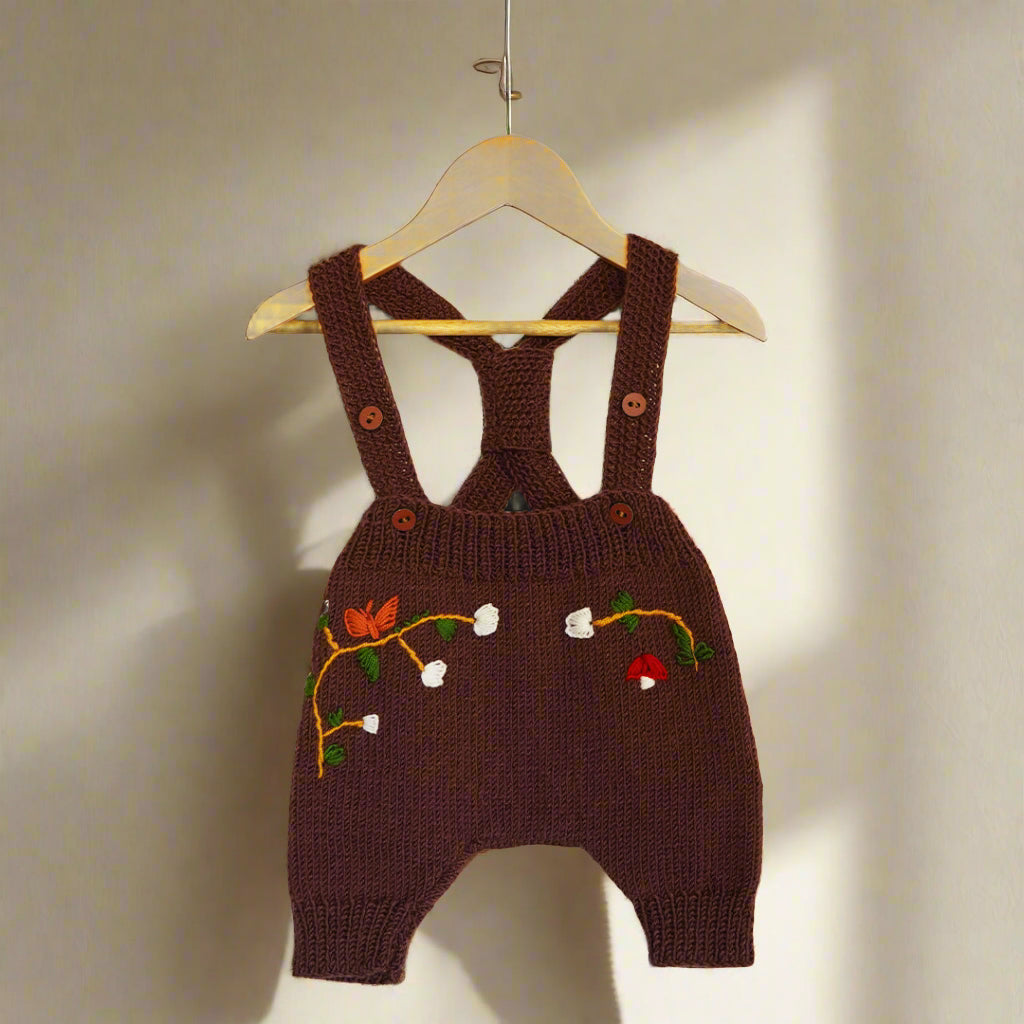  A beautifully hand-knitted baby romper in a rich chocolate brown color, featuring delicate hand-embroidered floral and nature-inspired details, including white blossoms, a butterfly, and a small red mushroom. The romper has adjustable cross-back straps with wooden button closures, adding a timeless, rustic charm. Made from soft yarn, it offers both warmth and comfort, perfect for little ones.