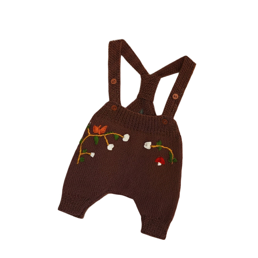  A beautifully hand-knitted baby romper in a rich chocolate brown color, featuring delicate hand-embroidered floral and nature-inspired details, including white blossoms, a butterfly, and a small red mushroom. The romper has adjustable cross-back straps with wooden button closures, adding a timeless, rustic charm. Made from soft yarn, it offers both warmth and comfort, perfect for little ones.