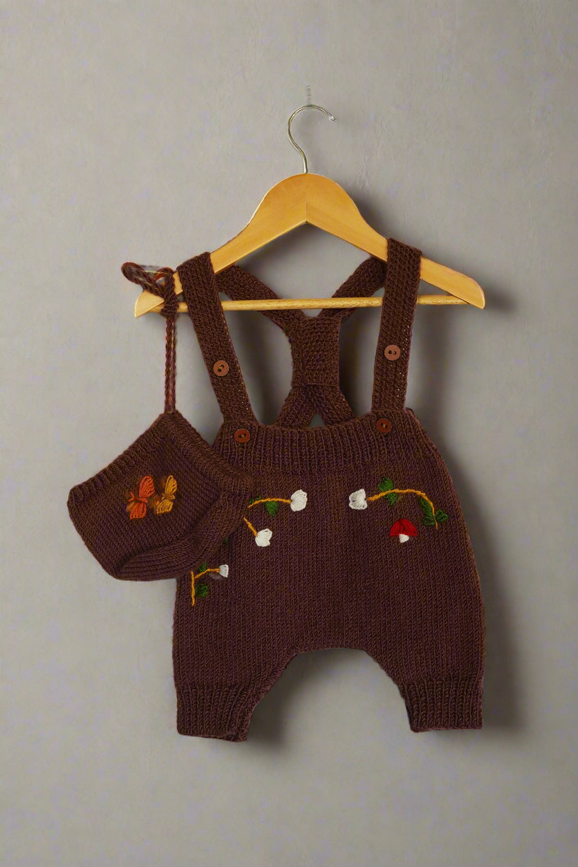 A beautifully hand-knitted baby romper in a rich chocolate brown color, featuring delicate hand-embroidered floral and nature-inspired details, including white blossoms, a butterfly, and a small red mushroom. The romper has adjustable cross-back straps with wooden button closures, adding a timeless, rustic charm. Made from soft yarn, it offers both warmth and comfort, perfect for little ones.