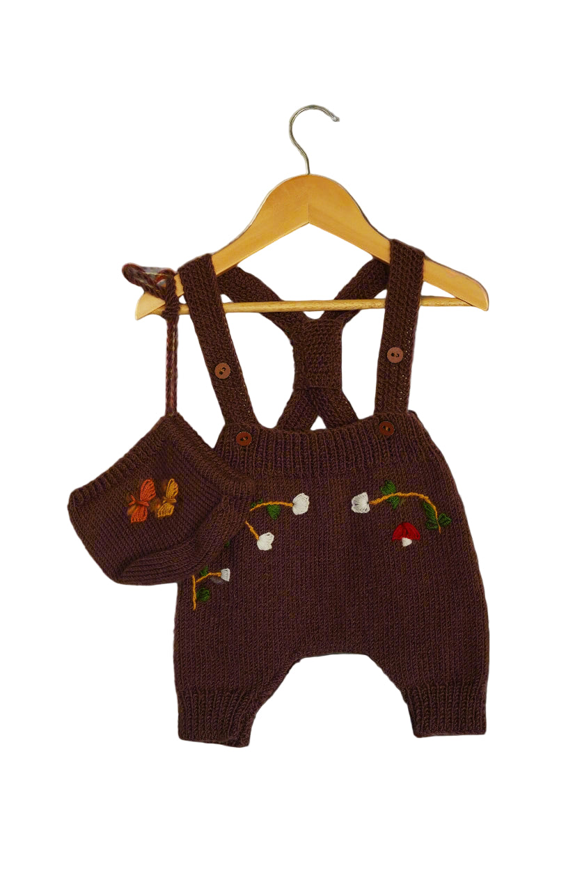 A beautifully hand-knitted baby romper in a rich chocolate brown color, featuring delicate hand-embroidered floral and nature-inspired details, including white blossoms, a butterfly, and a small red mushroom. The romper has adjustable cross-back straps with wooden button closures, adding a timeless, rustic charm. Made from soft yarn, it offers both warmth and comfort, perfect for little ones.