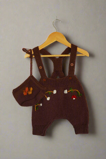 A beautifully hand-knitted baby romper in a rich chocolate brown color, featuring delicate hand-embroidered floral and nature-inspired details, including white blossoms, a butterfly, and a small red mushroom. The romper has adjustable cross-back straps with wooden button closures, adding a timeless, rustic charm. Made from soft yarn, it offers both warmth and comfort, perfect for little ones.
