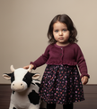 Young girl in a floral dress and purple cardigan holding a plush cow toy against a beige background