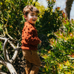 Child in a rust-colored sweater standing among green plants

