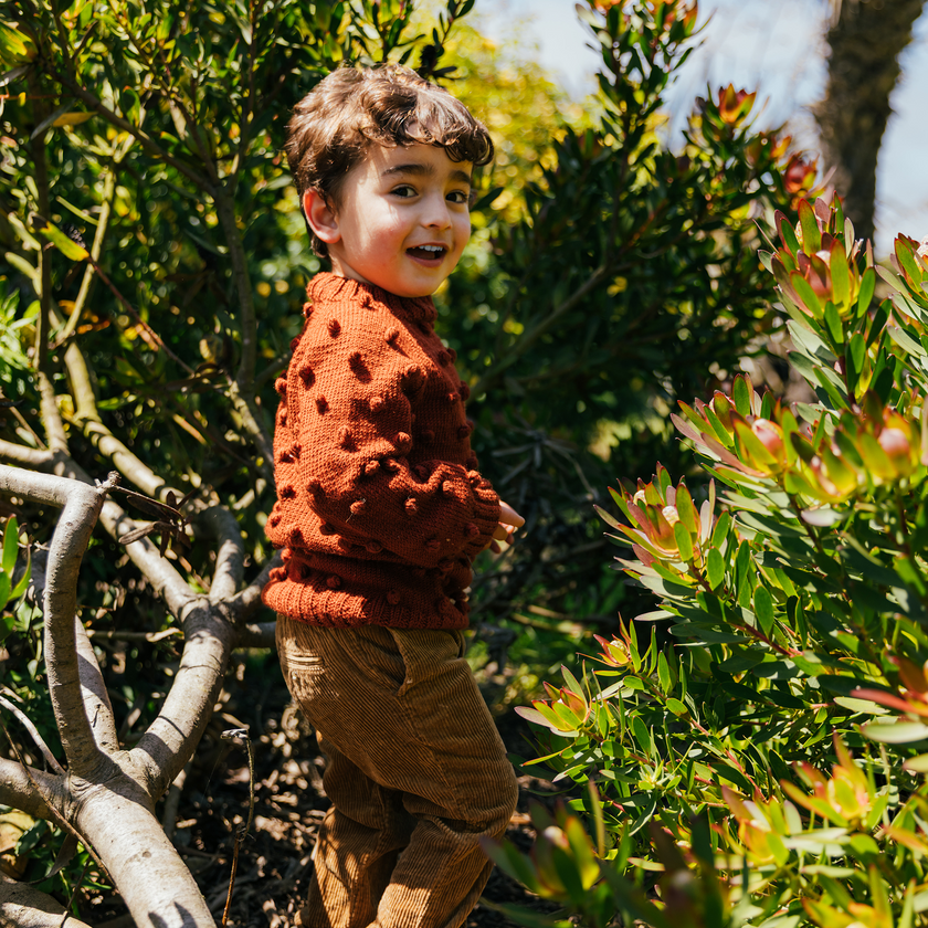 Child in a rust-colored sweater standing among green plants

