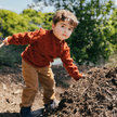Child in a rust sweater and beige pants standing on a pile of dirt with trees in the background